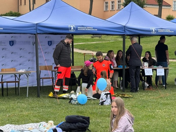 Obraz przedstawia wydarzenie plenerowe pod trzema przyległymi niebieskimi namiotami na trawie. Namioty należą do centrum naukowego. W środku kilka osób siedzi przy stołach, organizując lub uczestnicząc w wydarzeniu. Na pierwszym planie dwie osoby, jedna w czerwonym uniformie ratowniczym, zajmują się dziewczynką. Obok na trawie leżą niebieskie balony, a dziewczynka siedzi na kocu i obserwuje. W tle widać więcej osób, drzewa i kilka budynków.