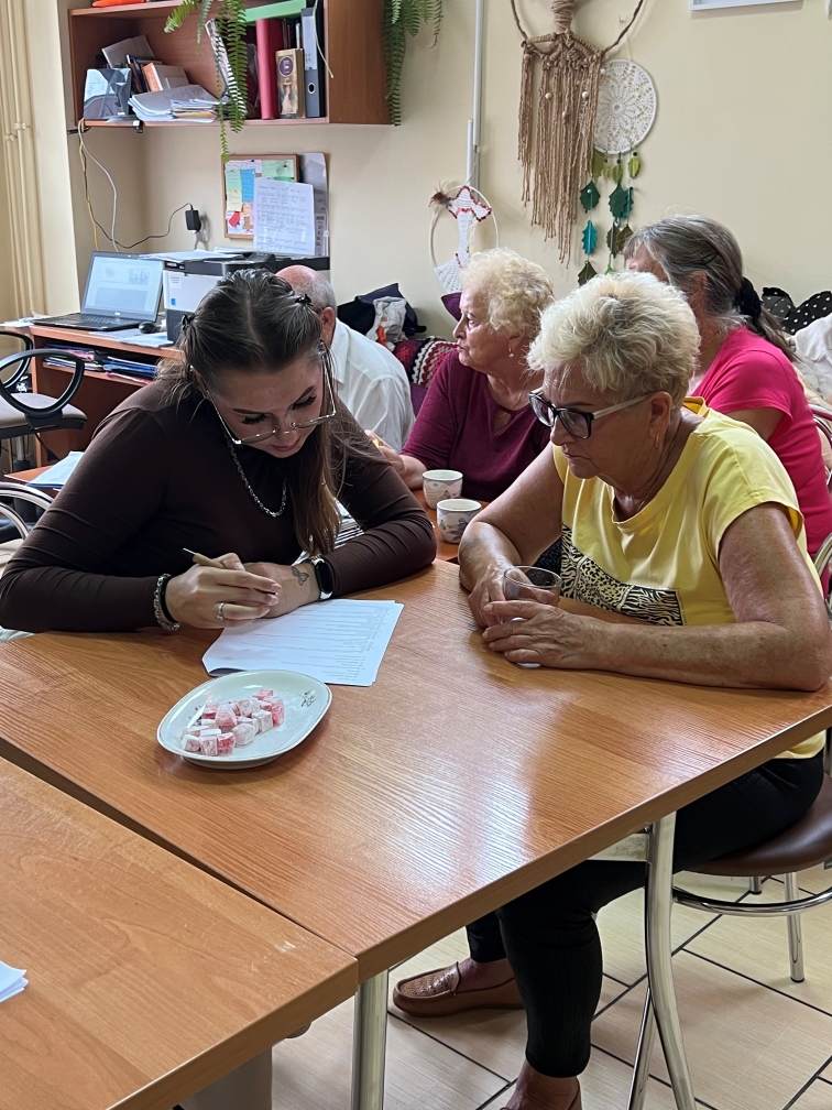 Obraz przedstawia grupę osób siedzących przy drewnianym stole w przytulnym pomieszczeniu przypominającym biuro lub salę spotkań. Na pierwszym planie kobieta pisze coś na kartce, a obok niej druga osoba uważnie się przygląda. Wokół stołu siedzi kilka osób, rozmawiających przy filiżankach kawy. W tle znajduje się ściana z dekoracyjnym makramowym elementem oraz regał wypełniony segregatorami, książkami i roślinami doniczkowymi. Na biurku za grupą stoi otwarty laptop.