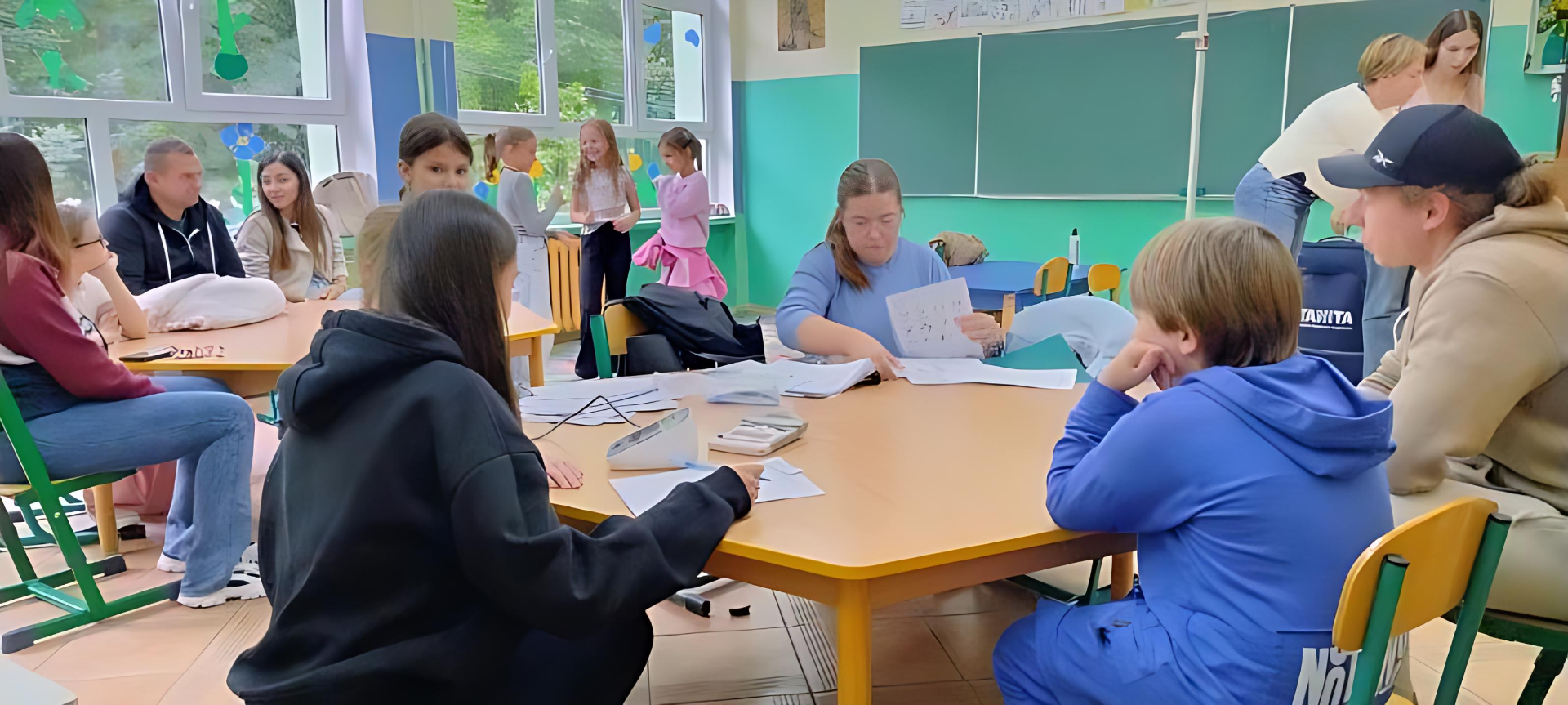 Obraz przedstawia salę lekcyjną, w której dzieci i dorośli biorą udział w różnych aktywnościach. Na pierwszym planie grupa dzieci i osoba dorosła siedzą przy okrągłym stole – część z nich przegląda kartki, inni uważnie się przyglądają. Po prawej stronie dziecko w niebieskim dresie siedzi tyłem do kamery, a obok niego inne, w czarnej bluzie, skupia się na pracy przy stole. Po lewej stronie dorosły w ciemnej kurtce i dżinsach siedzi obok kilkorga dzieci. W tle, po prawej stronie, znajduje się zielona tablica, a po lewej okna wpuszczające naturalne światło. Przy oknach kilka osób, w tym kobieta i dwoje dzieci, prowadzą rozmowę lub wykonują zadania.