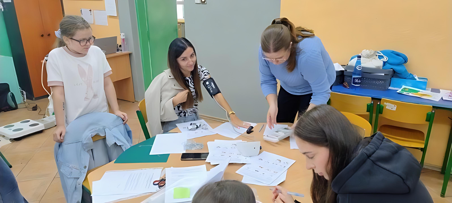 Obraz przedstawia pięć kobiet pracujących wspólnie przy jasnym, drewnianym stole w sali przypominającej klasę. Na stole leżą kartki, nożyczki i małe opakowanie. Jedna z kobiet, stojąca po prawej stronie w niebieskim swetrze, pochyla się nad materiałami. Naprzeciwko niej siedzi kobieta z długimi ciemnymi włosami, trzymając rękaw i angażując się w pracę. Po lewej stronie stoi kobieta w białej koszulce z wyblakowanym nadrukiem, obserwując działania. Dwie pozostałe osoby, widoczne od tyłu, są zajęte dokumentami lub pisaniem. W tle widać kolorowe ściany w odcieniach pomarańczowym i zielonym, a także otwarte półki i krzesła, co nadaje wnętrzu swobodną atmosferę.