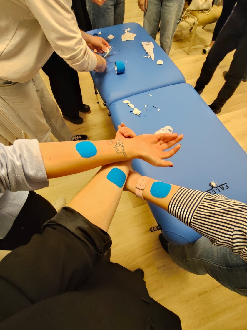 Na zdjęciu widać grupę osób zgromadzonych wokół stołu do masażu z niebieską powierzchnią. Na stole leżą kawałki taśmy oraz rolka niebieskiej taśmy kinezjologicznej. Jedna osoba przycina taśmę, a inne mają już fragmenty taśmy naklejone na przedramionach. Uczestnicy są ubrani swobodnie. Scena rozgrywa się prawdopodobnie w sali warsztatowej lub szkoleniowej – w tle widoczne są drewniana podłoga i krzesła.