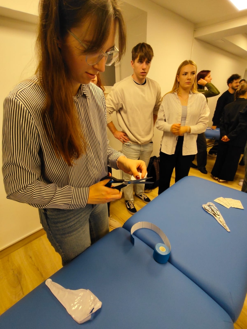 Na zdjęciu widać grupę osób zgromadzonych wokół niebieskiego stołu do masażu. Na pierwszym planie osoba w okularach i w paskowanej koszuli przycina nożyczkami niebieską taśmę. Na stole leży rolka taśmy, paski papieru oraz biały element przypominający rękawiczkę. W tle kilka osób w jasnych ubraniach obserwuje działania przy stole.