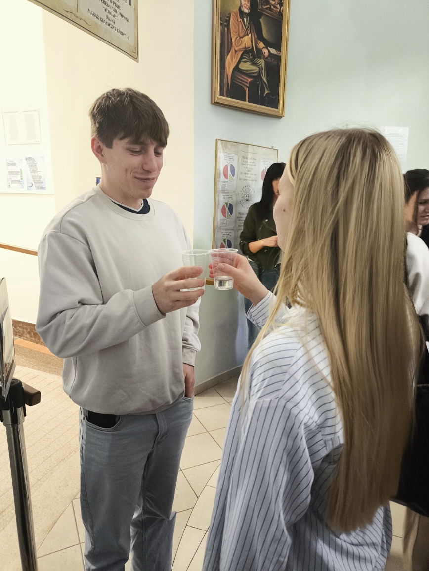 Na zdjęciu widać młodego mężczyznę i kobietę stojących w pomieszczeniu i stukających się plastikowymi kubkami. Mężczyzna lekko się uśmiecha i trzyma kubek w prawej ręce, a lewą ma w kieszeni. Kobieta stoi tyłem do aparatu. Znajdują się na podłodze z płytek, a w tle widoczne są obraz w ramie, plakat z wykresami oraz osoba korzystająca z telefonu.