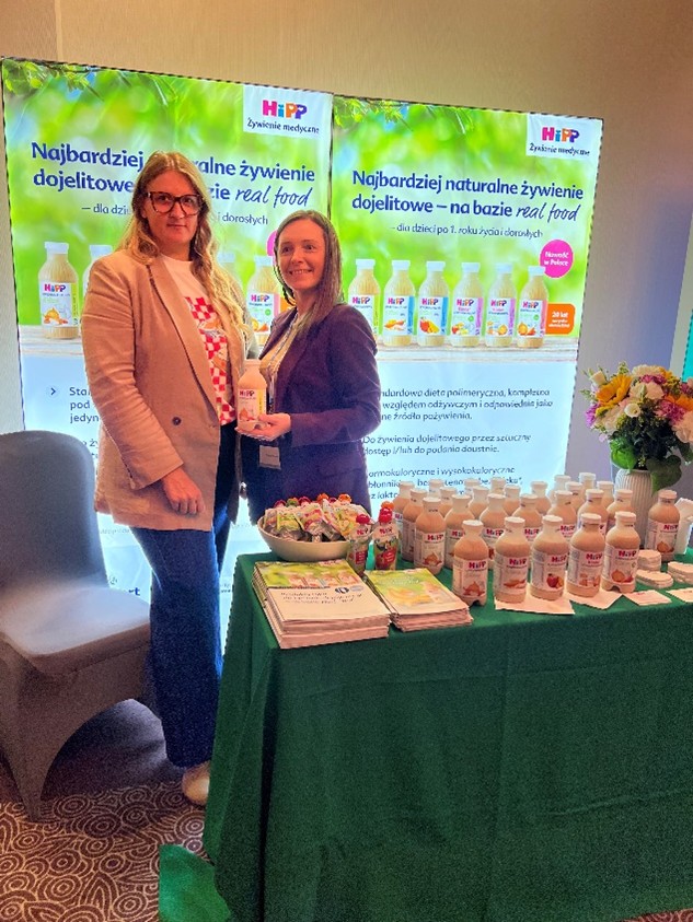 Na zdjęciu widać dwie kobiety stojące przy stoliku wystawowym podczas targów lub wydarzenia. Za nimi znajdują się dwa podświetlane banery z logo marki HiPP oraz grafikami przedstawiającymi produkty. Na stoliku, przykrytym zielonym obrusem, ustawiono butelki produktów HiPP, ulotki informacyjne oraz miseczkę z przekąskami. Z boku znajduje się również bukiet kwiatów.