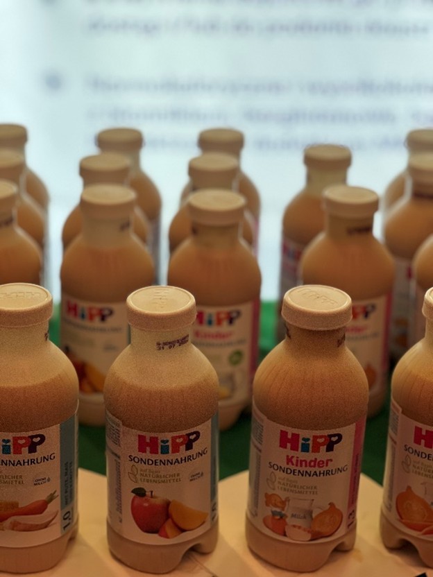 Na zdjęciu widać kilka jasnobrązowych butelek z beżowymi zakrętkami, ustawionych blisko siebie w rzędach. Na etykietach widnieje logo marki HiPP oraz kolorowe grafiki owoców, takich jak jabłka i banany.