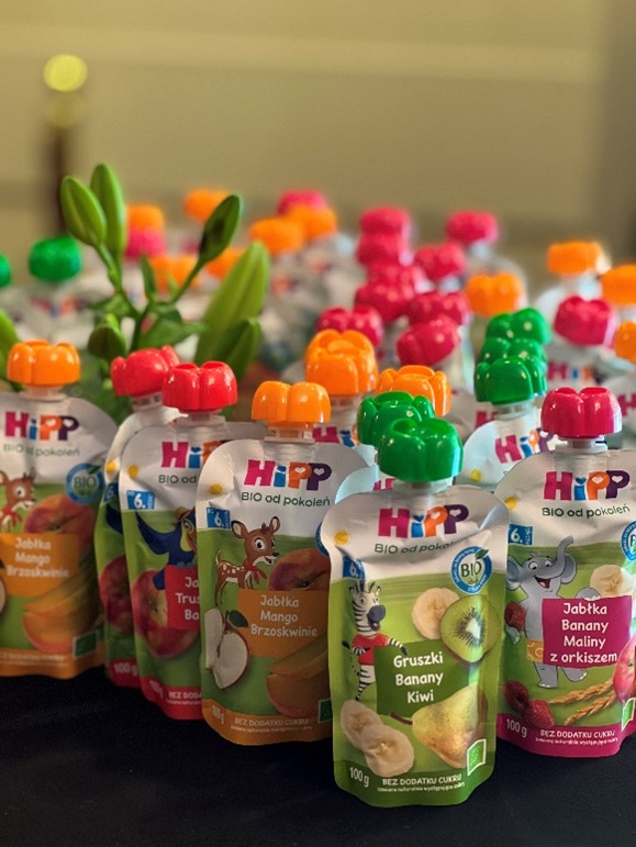 Na zdjęciu widać kolorowe saszetki z owocowymi musami, ustawione blisko siebie na ciemnej powierzchni. Każda saszetka ma wesoły design z ilustracjami owoców i zwierząt, a zakrętki w żywych kolorach – m.in. pomarańczowym, zielonym, różowym i czerwonym. W tle widoczne są zielone liście, dodające naturalnego akcentu. Etykiety zawierają grafiki owoców i zwierząt oraz informacje o smakach.