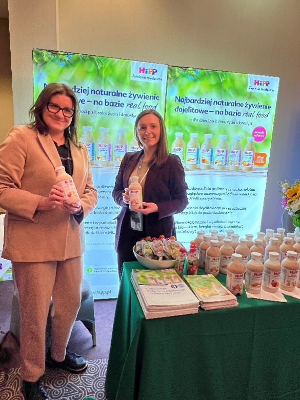 Na zdjęciu widać dwie osoby stojące przy stanowisku promocyjnym marki HiPP. Trzymają butelki z widocznym logo HiPP. Za nimi znajdują się dwa pionowe banery z zielonym, roślinnym motywem, prezentujące logo i produkty marki. Przed banerami ustawiono stolik przykryty zielonym obrusem, na którym znajdują się butelki, saszetki z musami w misce oraz stosy broszur. Na brzegu stołu stoi bukiet kwiatów jako element dekoracyjny.