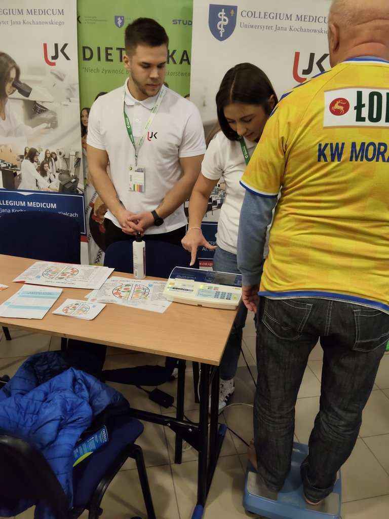 Na zdjęciu widać stoisko eventowe, na którym prowadzone są badania zdrowotne. Po lewej stronie dwie osoby w koszulkach z logo „UJK” pracują przy stoliku, omawiając dane z urządzenia i prezentując materiały informacyjne. Na stole leżą ulotki oraz cyfrowa waga. Po prawej stronie uczestnik wydarzenia stoi na niebieskiej wadze. W tle znajdują się banery „Collegium Medicum Uniwersytetu Jana Kochanowskiego” oraz „Dietetyka.”