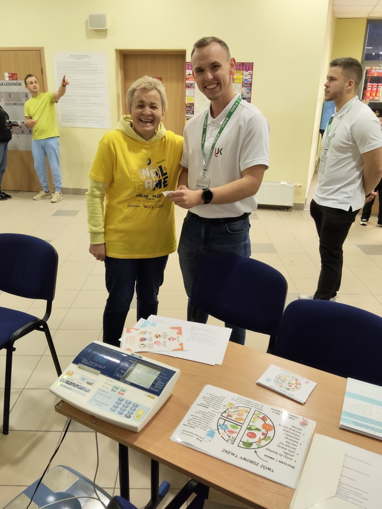 Zdjęcie przedstawia pomieszczenie, w którym znajdują się osoby. Na pierwszym planie stoi stół z elektronicznym urządzeniem oraz kilkoma kartami z diagramami, prawdopodobnie o tematyce żywieniowej. Za stołem dwie osoby w żółtej i białej koszulce rozmawiają ze sobą w przyjaznej atmosferze. W tle kolejna osoba w żółtej koszulce wskazuje coś poza kadrem, a obok niej stoi ktoś w białej koszulce z identyfikatorem. W pomieszczeniu widać jasne ściany, płytki na podłodze oraz tablicę informacyjną z plakatami.