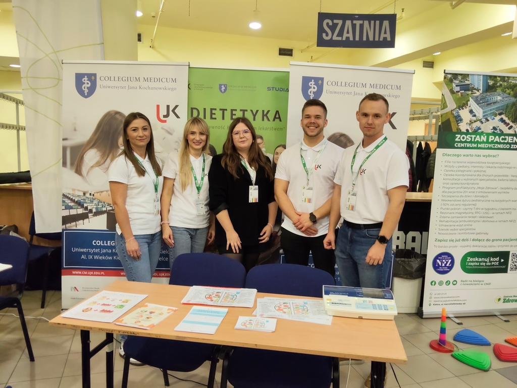 Zdjęcie przedstawia pięć osób stojących za stołem podczas wydarzenia lub wystawy. Za nimi widoczne są banery związane z tematyką medyczną i edukacyjną, m.in. „Collegium Medicum”. Osoby mają na sobie białe koszulki i identyfikatory. Na stole leżą kolorowe materiały informacyjne. Nad grupą znajduje się tabliczka z napisem „SZATNIA”.