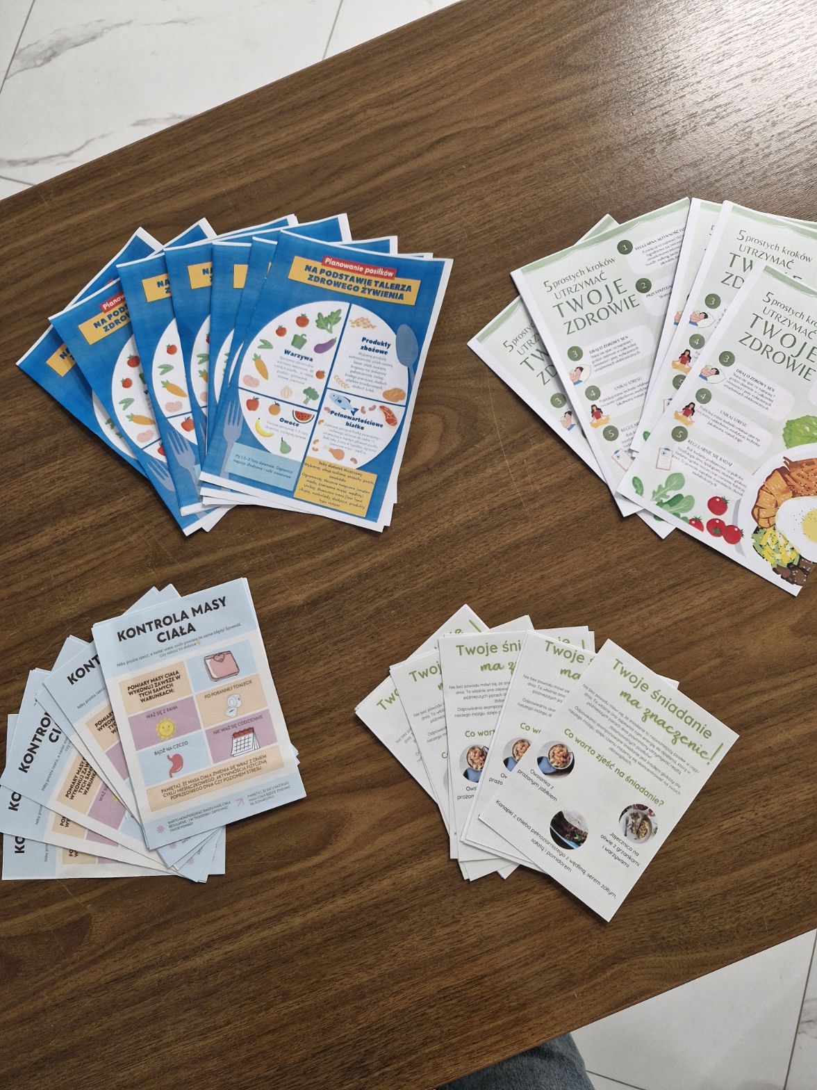 Na zdjęciu widać drewniany stół, na którym leżą cztery stosy ulotek. W lewym górnym rogu znajdują się niebieskie ulotki z kolorowym wykresem dotyczącym odżywiania, w prawym górnym – jasnozielone i białe z ilustracjami jedzenia oraz wskazówkami zdrowotnymi. W lewym dolnym rogu widać jasnoniebieskie broszury poświęcone kontroli wagi, a w prawym dolnym – białe ulotki z zielonym tekstem o śniadaniach i poradach żywieniowych. Pod stołem widać podłogę z dużych białych płytek z delikatnym szarym wzorem.