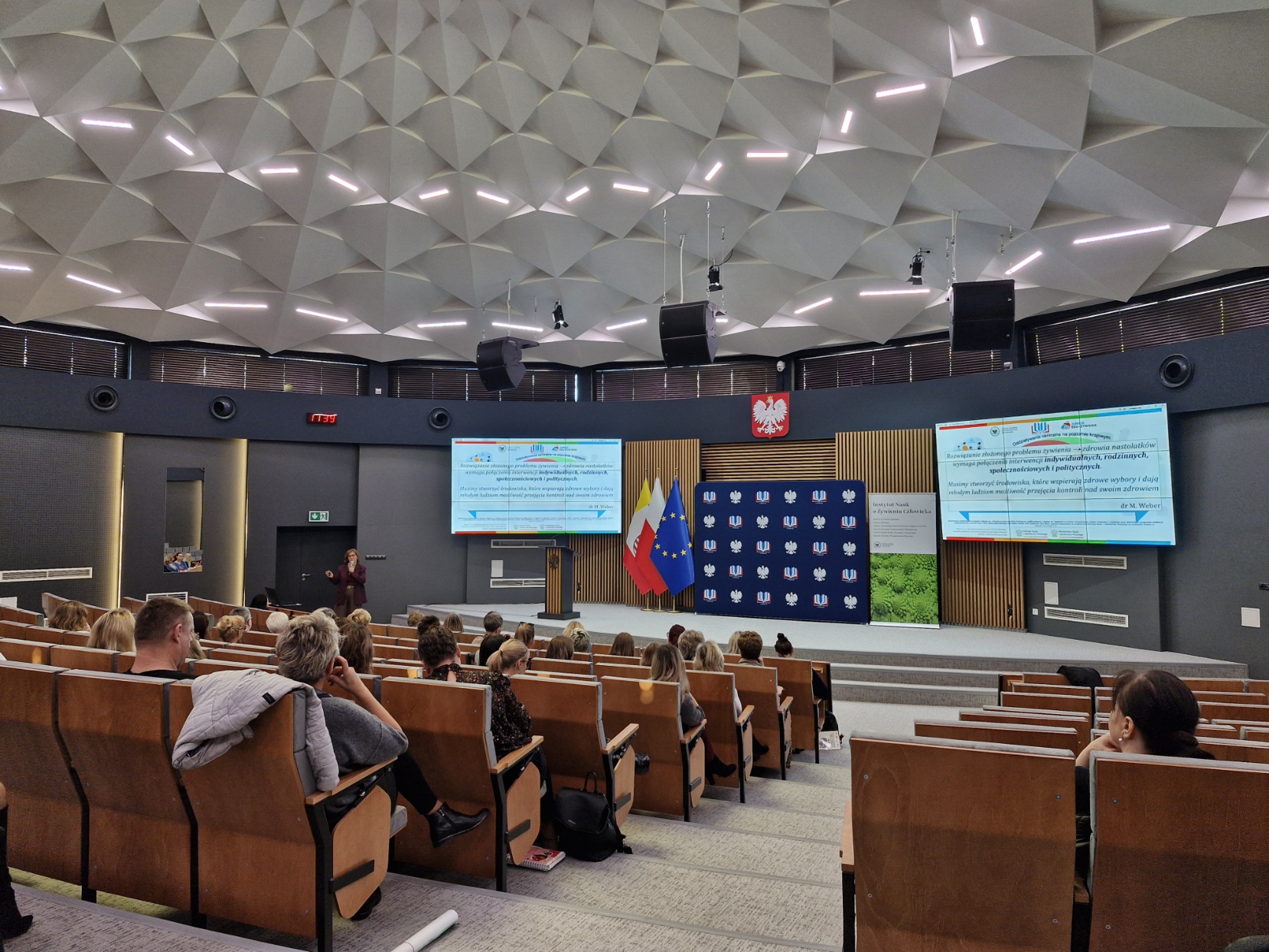 Na zdjęciu widać nowoczesną salę audytoryjną wypełnioną publicznością oglądającą prezentację. Pomieszczenie ma geometryczny, podświetlany sufit o ciekawej strukturze. Uczestnicy siedzą w rzędach drewnianych krzeseł z ciemnymi podłokietnikami, a uwaga skupia się na scenie z przodu. Na dwóch dużych ekranach wyświetlana jest prezentacja z tekstem. Za mównicą znajduje się tło z powtarzającym się logo, a po lewej stronie sceny przemawia prelegent. Ściany ozdobione są pionowymi drewnianymi panelami oraz dużym emblematem w czerwono-białych barwach.