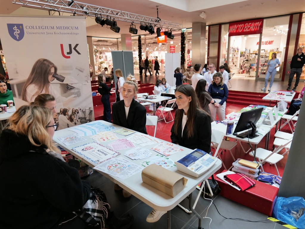 Obraz przedstawia wydarzenie odbywające się w centrum handlowym. Na pierwszym planie cztery osoby rozmawiają przy stole z kolorowymi ulotkami. Za nimi widoczny jest baner Collegium Medicum Uniwersytetu Jana Kochanowskiego z ilustracją mikroskopu. Obok znajduje się kolejne stanowisko z monitorem i materiałami informacyjnymi. W tle przechodzą osoby, a na sklepach widoczne są m.in. szyldy ‘WAY TO BEAUTY’. Podłoga ma intensywny czerwony kolor, a nad strefą zawieszono oświetlenie i nagłośnienie.