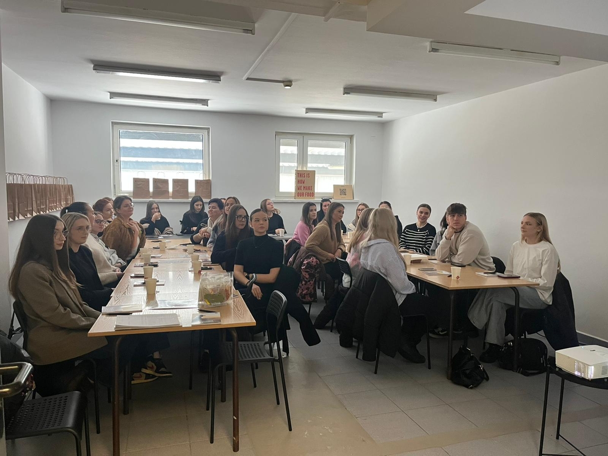 Obraz przedstawia grupę osób siedzących przy długim prostokątnym stole w jasnym pomieszczeniu z białymi ścianami i dwoma oknami. Na stole znajdują się kubki, notatniki i szklany słoik. W tle widać proste dekoracje, takie jak plakaty na parapetach i papierowe torby na bocznej półce. Grupa prowadzi dyskusję lub spotkanie.