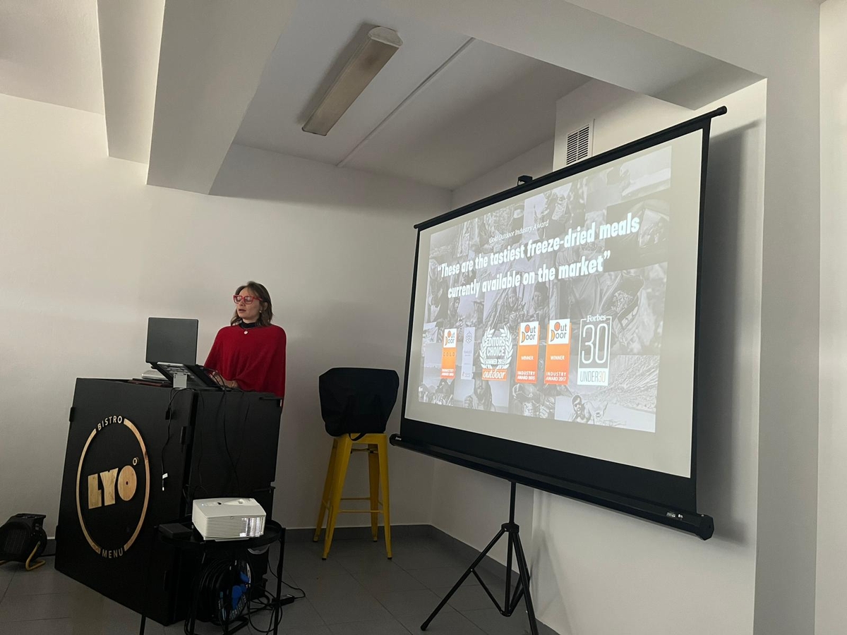 Obraz przedstawia ustawienie do prezentacji w pomieszczeniu. Po lewej stronie osoba stoi za czarną mównicą z okrągłym logo „LYO Bistro Menu” i korzysta z laptopa. Na podłodze przed mównicą znajduje się projektor z widocznymi kablami. Po prawej stronie duży ekran projekcyjny wyświetla slajd przedstawiający liczne nagrody związane z daniami liofilizowanymi. Białe ściany i sufit tworzą jasne, uporządkowane otoczenie.