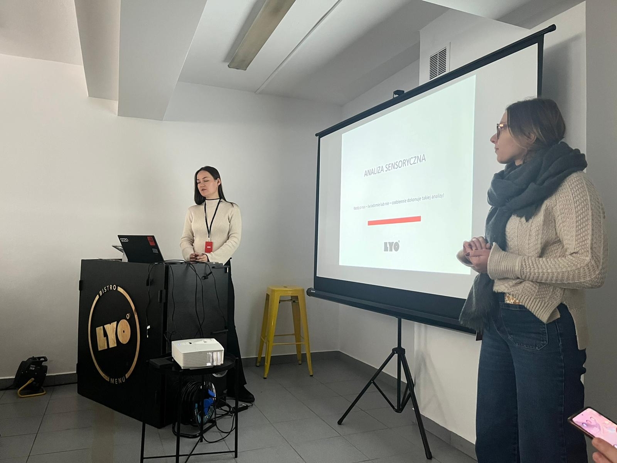 Obraz przedstawia prezentację w pomieszczeniu z dwiema osobami. Jedna stoi za czarną mównicą z logo „LYO”, obok laptopa i podłączonych kabli, w tym przewodu do projektora. Druga znajduje się przy białym ekranie projekcyjnym, na którym wyświetlony jest slajd po polsku. W tle widoczny jest żółty stołek.