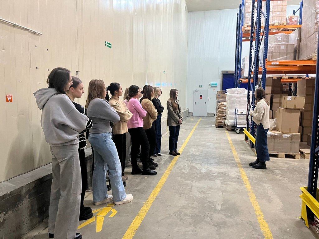 Obraz przedstawia dziewięć osób stojących w magazynie, ustawionych wzdłuż lewej strony i słuchających osoby przemawiającej z prawej. Wysokie, jasne ściany i regały wypełnione kartonami tworzą uporządkowaną przestrzeń. Betonowa podłoga ma żółte oznaczenia, a w tle widać wózek z torbami. Oświetlenie jest jasne i równomierne.