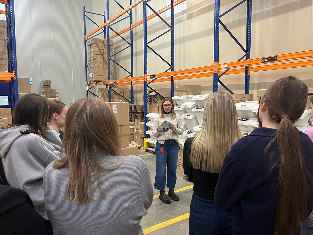 Obraz przedstawia grupę osób w magazynie, zgromadzonych wokół prowadzącej, która trzyma kartkę i omawia jakiś temat. Uczestnicy stoją tyłem do kamery. W magazynie widać wysokie niebieskie regały z pomarańczowymi belkami, wypełnione kartonami i białymi pakunkami. Podłoga jest betonowa z żółtymi oznaczeniami, a otoczenie ma industrialny charakter z wysokim sufitem i neutralnymi ścianami.