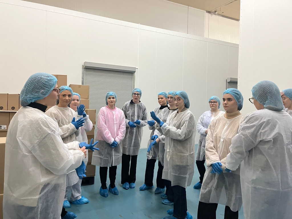 Obraz przedstawia grupę osób w sterylnym, kontrolowanym pomieszczeniu, prawdopodobnie w laboratorium lub zakładzie przetwórstwa żywności. Uczestnicy mają na sobie siatki ochronne, jednorazowe fartuchy, ochraniacze na obuwie i rękawice. Jasne, białe ściany i niebieska podłoga podkreślają sterylny charakter miejsca. Z tyłu znajduje się metalowa roleta oraz stosy kartonów. Grupa wygląda na zaangażowaną w rozmowę lub instruktaż, zwracając uwagę na osobę mówiącą po lewej stronie.