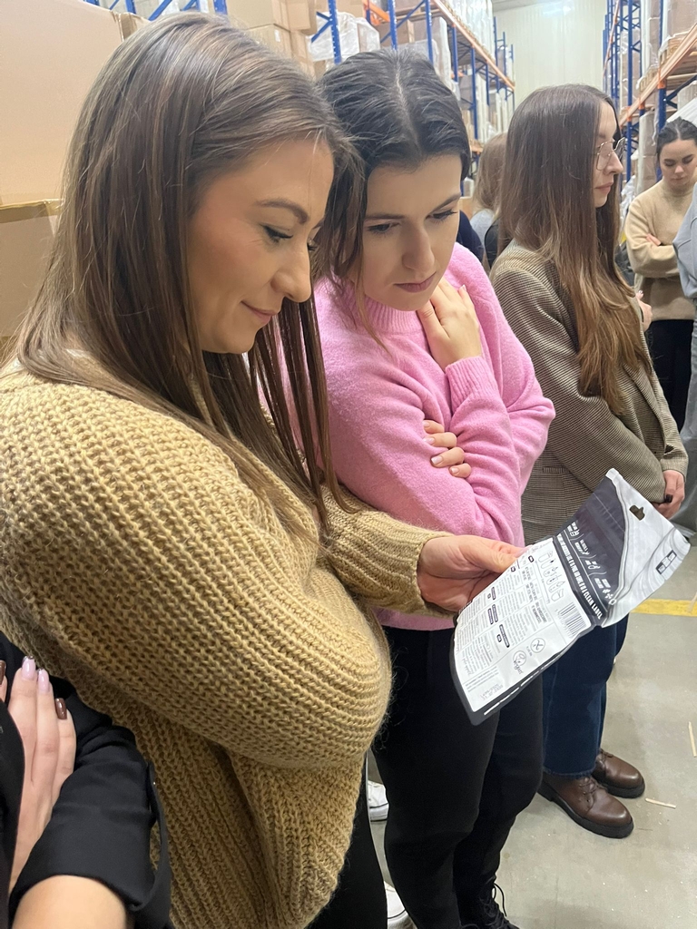 Obraz przedstawia grupę kobiet stojących w alejce magazynowej. Dwie osoby na pierwszym planie oglądają zapakowany produkt. Wysokie regały po obu stronach wypełnione są pudełkami. Pozostałe uczestniczki stoją w pobliżu i również przyglądają się produktowi lub rozmawiają. Jasne oświetlenie podkreśla przemysłowy charakter przestrzeni z metalowymi regałami i kartonami.