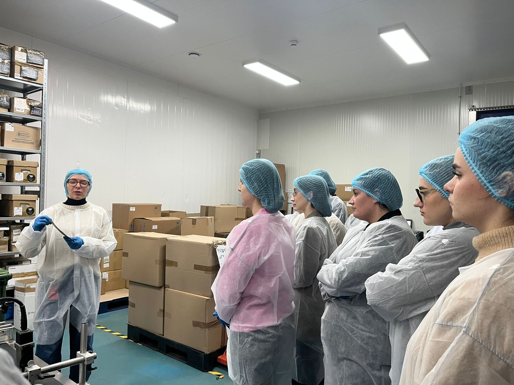 Obraz przedstawia grupę osób w pomieszczeniu magazynowym, prawdopodobnie związanym z obsługą żywności lub materiałów laboratoryjnych. W jasnym, dobrze oświetlonym wnętrzu uczestnicy w siatkach ochronnych i fartuchach stoją wokół osoby prowadzącej pokaz lub omówienie. Prowadzący trzyma przedmiot, którego nie widać wyraźnie. W tle znajdują się regały z opisanymi pudełkami oraz stosy kartonów przy ścianie.