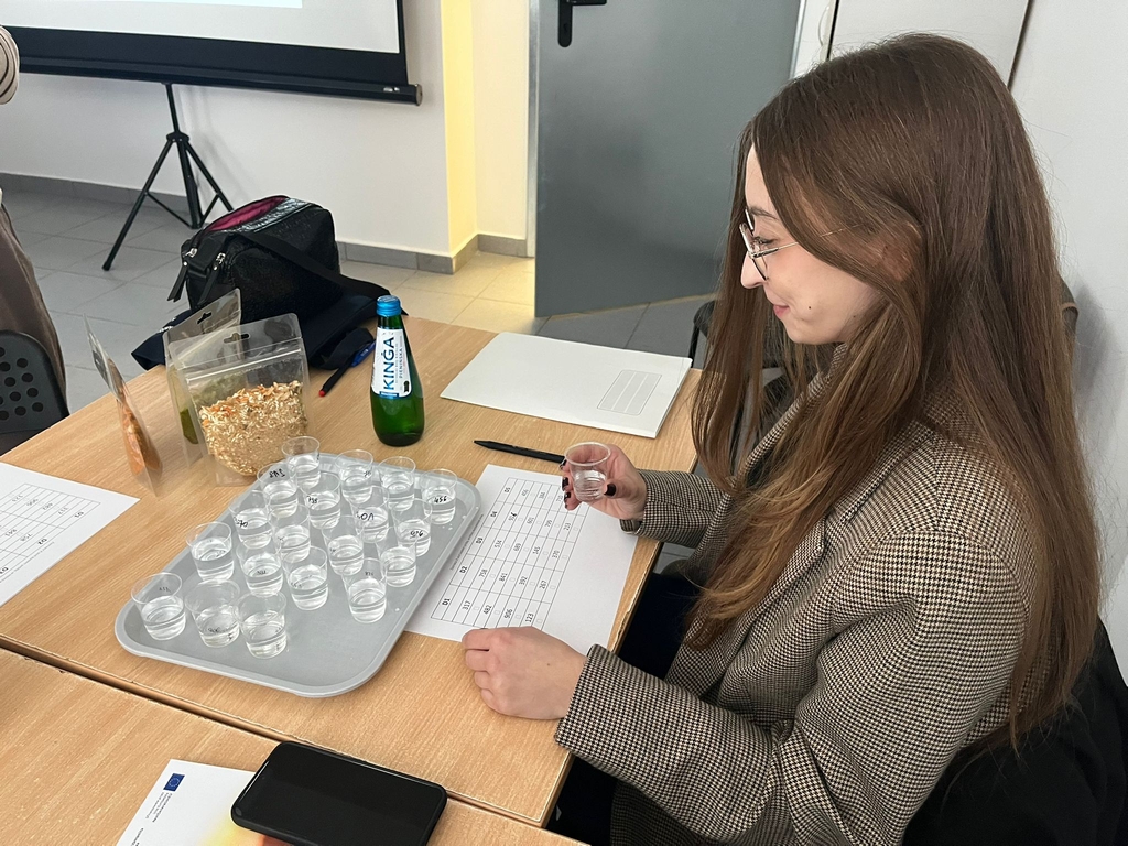 Obraz przedstawia osobę siedzącą przy drewnianym stole. Trzyma mały plastikowy kubek z tacki stojącej przed nią, na której znajduje się więcej takich kubków. Na stole leżą arkusze z tabelami, długopis, smartfon, butelka wody „KINGA” oraz strunowy worek z suszoną żywnością. W tle widać ekran projektora i częściowo torbę na krześle.