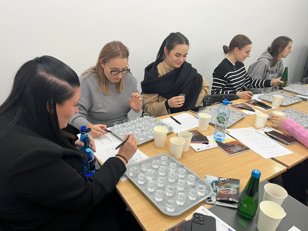 Obraz przedstawia sześć osób siedzących przy prostokątnym drewnianym stole w jasnym pomieszczeniu. Grupa pracuje z tackami wypełnionymi małymi plastikowymi kubkami i uzupełnia arkusze z wydrukowanymi tabelami. Na stole leżą tacki, jednorazowe kubki, butelki wody i broszury. Otoczenie jest proste, z białymi ścianami.