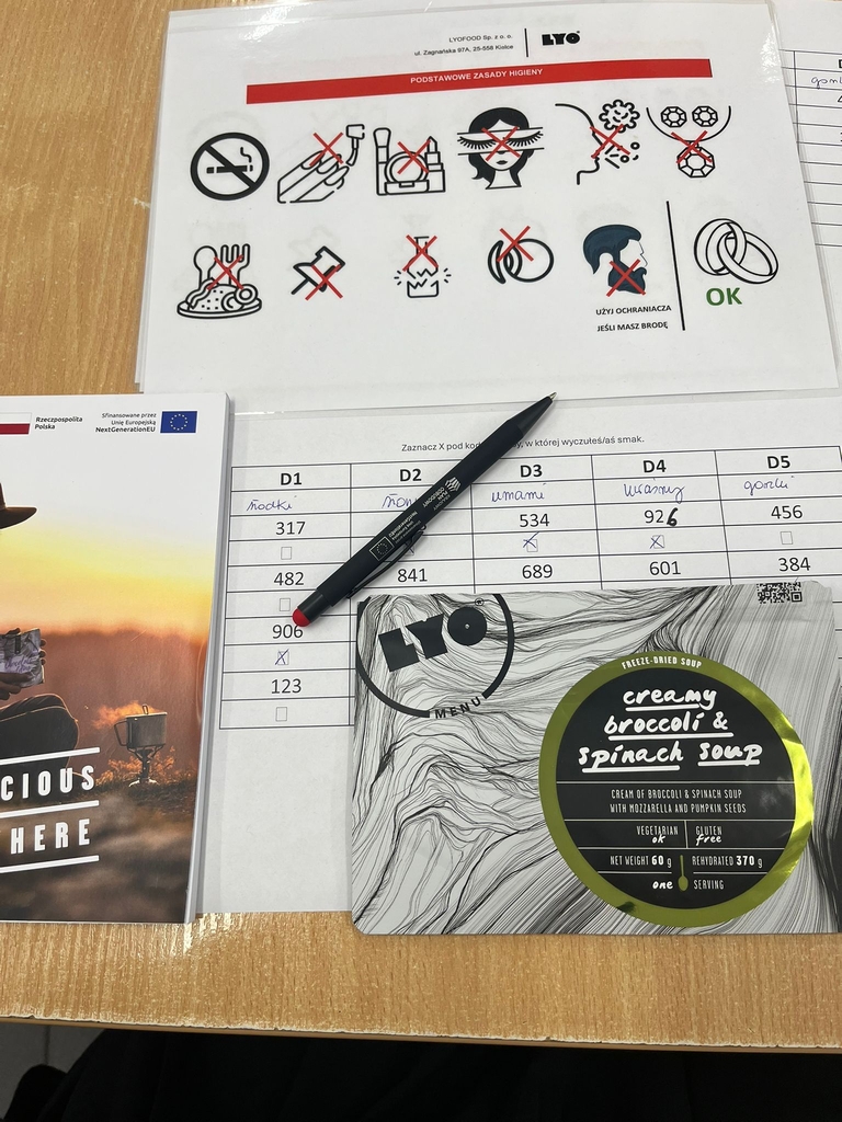 Na jasnym drewnianym blacie leżą różne przedmioty. W lewym górnym rogu znajduje się zalaminowana karta „Podstawowe Zasady Higieny” z ikonami i czerwonymi znakami X oraz logo Lyo i napisem „Użyj ochraniacza jeśli masz brodę”. Poniżej leży długopis na wypełnionej tabeli. Obok widać fragment broszury z napisem „CONSCIOUS”. W prawym dolnym rogu znajduje się opakowanie liofilizowanej zupy Lyo Menu „creamy broccoli & spinach soup”, oznaczone m.in. jako wegetariańskie i bezglutenowe.