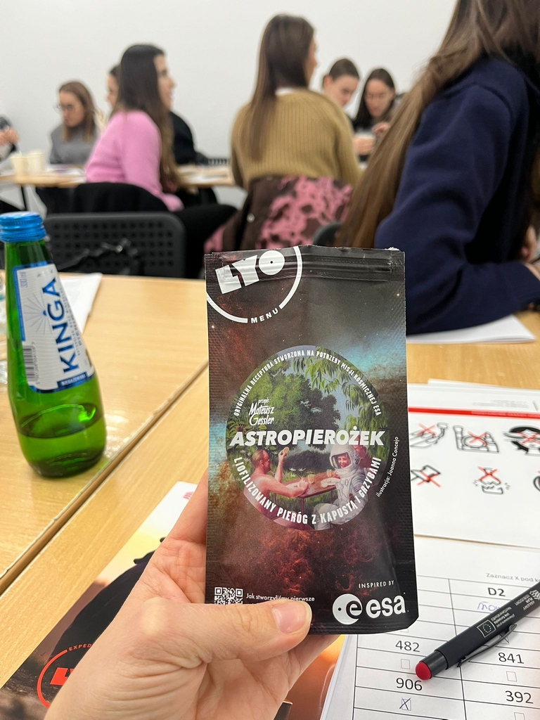 Obraz przedstawia salę lekcyjną z osobami siedzącymi przy stołach w tle. Na pierwszym planie widać paczkę produktu „ASTROPIEROŻEK” z kolorową grafiką oraz małą butelkę wody „KINGA”. Na stole leżą także długopis i dokumenty z tabelami i tekstem.