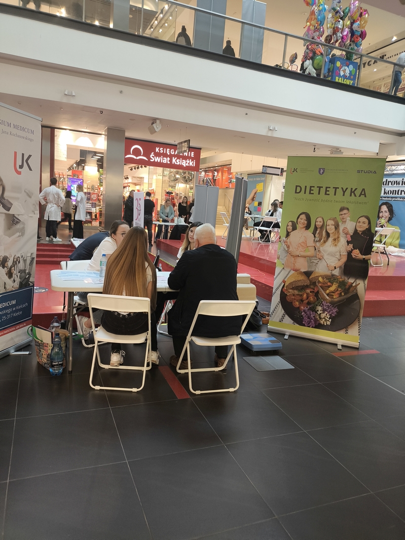 Obraz przedstawia wydarzenie w nowoczesnej przestrzeni wewnętrznej, prawdopodobnie w centrum handlowym lub hali wystawowej. Na pierwszym planie dwie osoby siedzą przy stole z materiałami i dokumentami. Po obu stronach widoczne są banery — jeden związany z instytucją medyczną lub edukacyjną, drugi z dietetyką. W tle znajduje się sklep ‘Świat Książki’, przechodnie oraz kolorowe balony, co sugeruje festynowy lub promocyjny charakter wydarzenia.