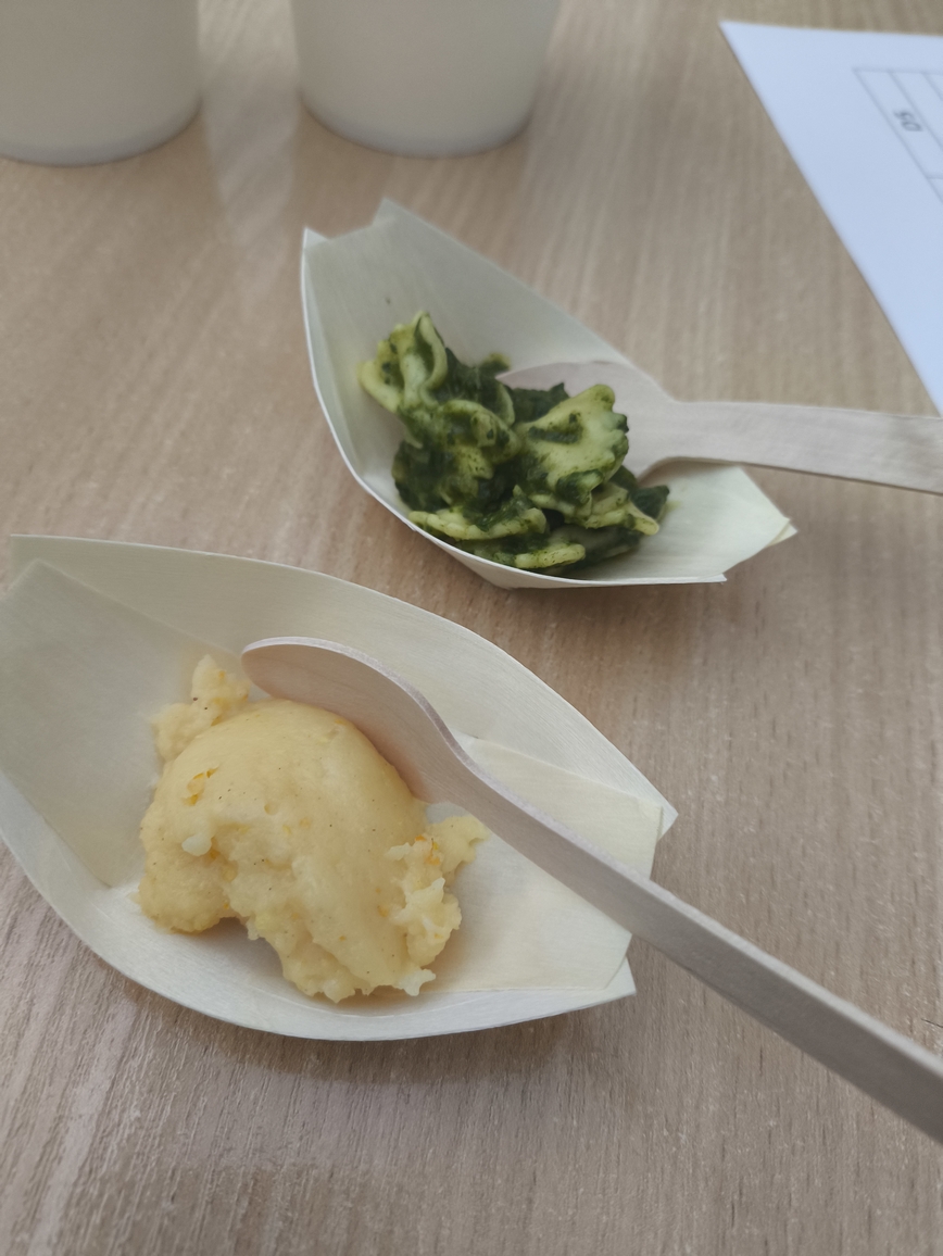 Obraz przedstawia dwa małe, drewniane naczynia w kształcie łódek na jasnym drewnianym stole. Jedno zawiera puree lub podobną kremową potrawę, a drugie makaron z zielonym sosem pesto. Na każdym naczyniu leży mała drewniana łyżeczka. W rogu widać fragment białej kartki z tekstem.