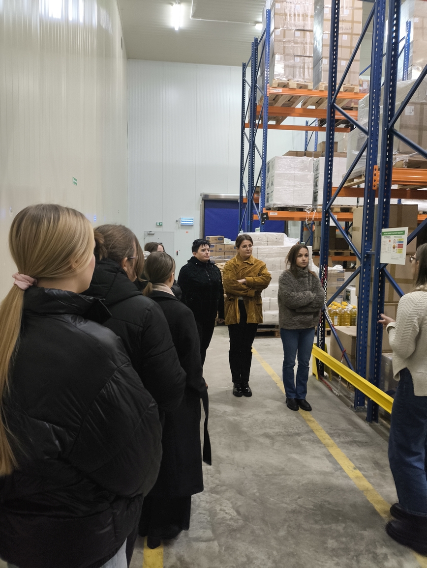 Obraz przedstawia grupę osób stojących w magazynie lub hali magazynowej z wysokim sufitem. Po prawej stronie widać metalowe regały wypełnione kartonami, niektóre owinięte folią. Osoby stoją w linii, zwracając uwagę na osobę przemawiającą. Podłoga betonowa ma żółte oznaczenia bezpieczeństwa.