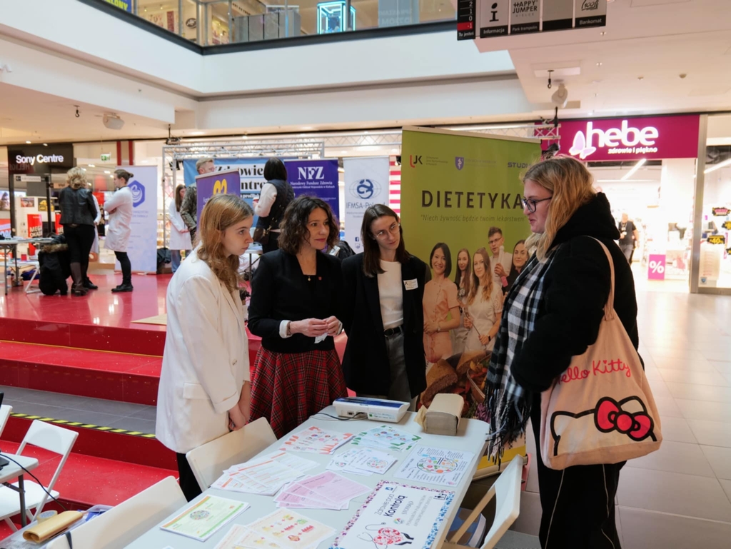 Obraz przedstawia grupę osób rozmawiających podczas wydarzenia w przestrzeni publicznej, prawdopodobnie targów zdrowotnych lub dietetycznych. Na pierwszym planie kilka osób stoi przy stole z ulotkami i materiałami informacyjnymi. W tle widoczny jest duży baner związany z dietetyką. Miejsce jest dobrze oświetlone, z czerwonym podłożem i nowoczesnym wystrojem.