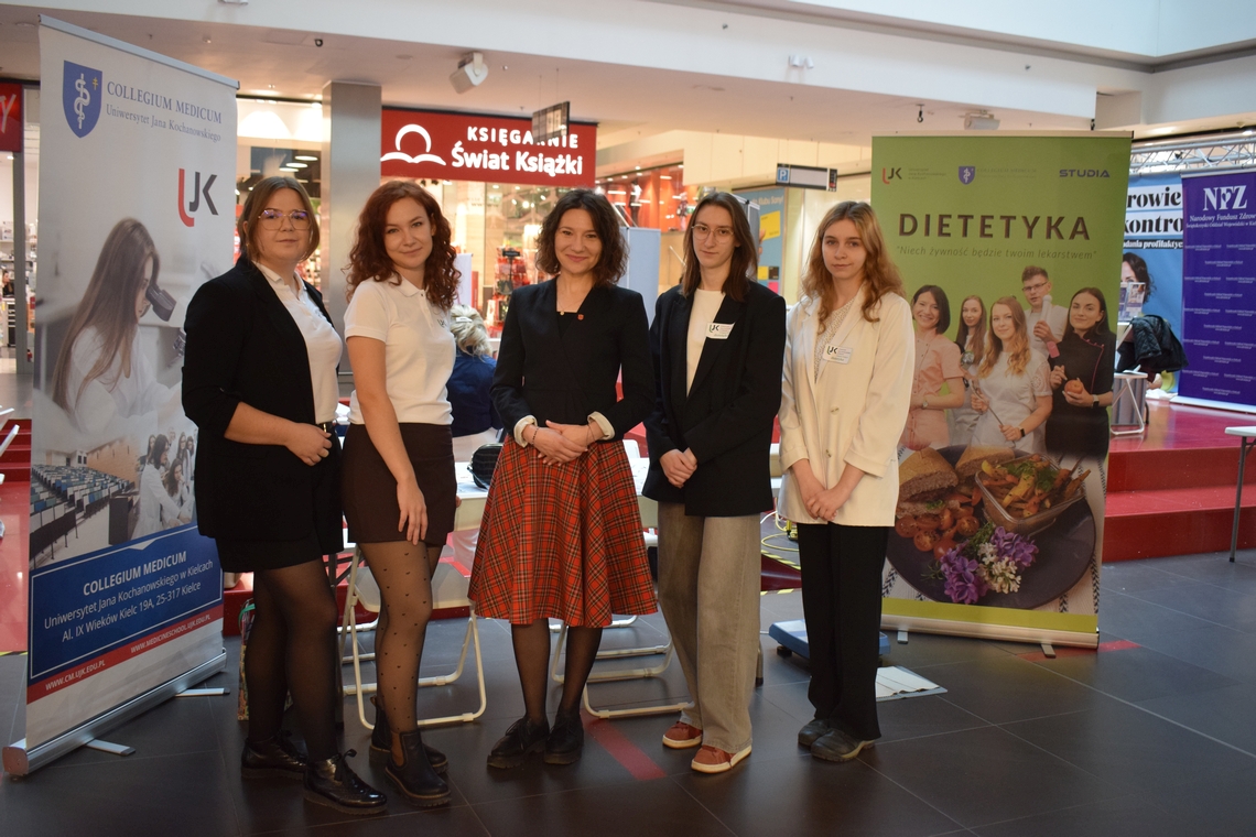 Obraz przedstawia pięć osób stojących przed banerami informacyjnymi w przestrzeni wewnętrznej, prawdopodobnie podczas wydarzenia. Po lewej stronie znajduje się baner Collegium Medicum, a po prawej baner Dietetyka. W tle widać kolorowe oznaczenia i stoiska, m.in. księgarnię oraz materiały związane ze zdrowiem.