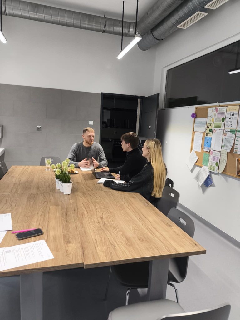 Obraz przedstawia spotkanie biznesowe w nowoczesnym biurze. Trzy osoby siedzą przy dużym, prostokątnym stole, rozmawiając ze sobą. Na stole stoją doniczki z roślinami. W pomieszczeniu widać tablicę z notatkami i dokumentami oraz sufit z widocznymi instalacjami i oświetleniem.