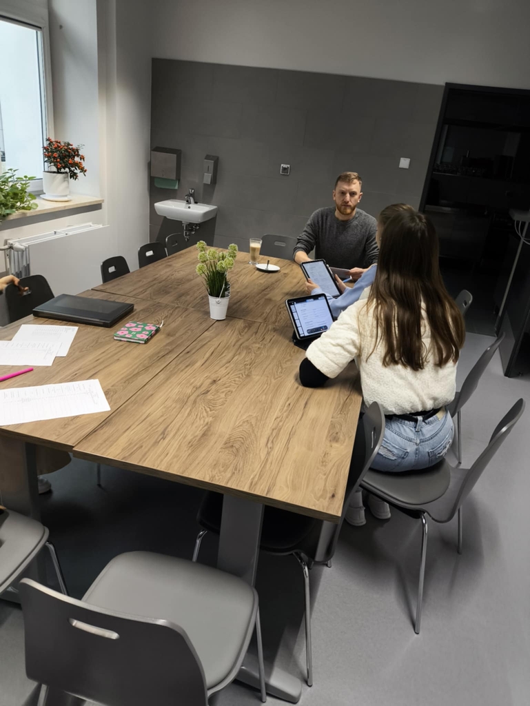 Obraz przedstawia nowoczesną salę konferencyjną z prostokątnym drewnianym stołem otoczonym szarymi krzesłami. Przy stole siedzą dwie osoby, obie korzystają z tabletów. Na stole leżą dokumenty, zamknięty laptop i dwie małe rośliny w wazonach. W pomieszczeniu widać okno z roślinami na parapecie oraz umywalkę z dozownikiem na tylnej ścianie. Jasne światło dzienne oświetla wnętrze.