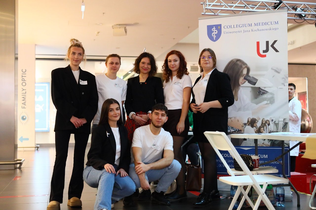 Obraz przedstawia siedem osób pozujących do wspólnego zdjęcia w przestrzeni wystawowej. W tle znajduje się duży baner COLLEGIUM MEDICUM Uniwersytet Jana Kochanowskiego z ilustracjami przedstawiającymi pracę studentów. Otoczenie jest dobrze oświetlone, z widoczną kafelkową podłogą i częściowo widocznymi dodatkowymi oznaczeniami.
