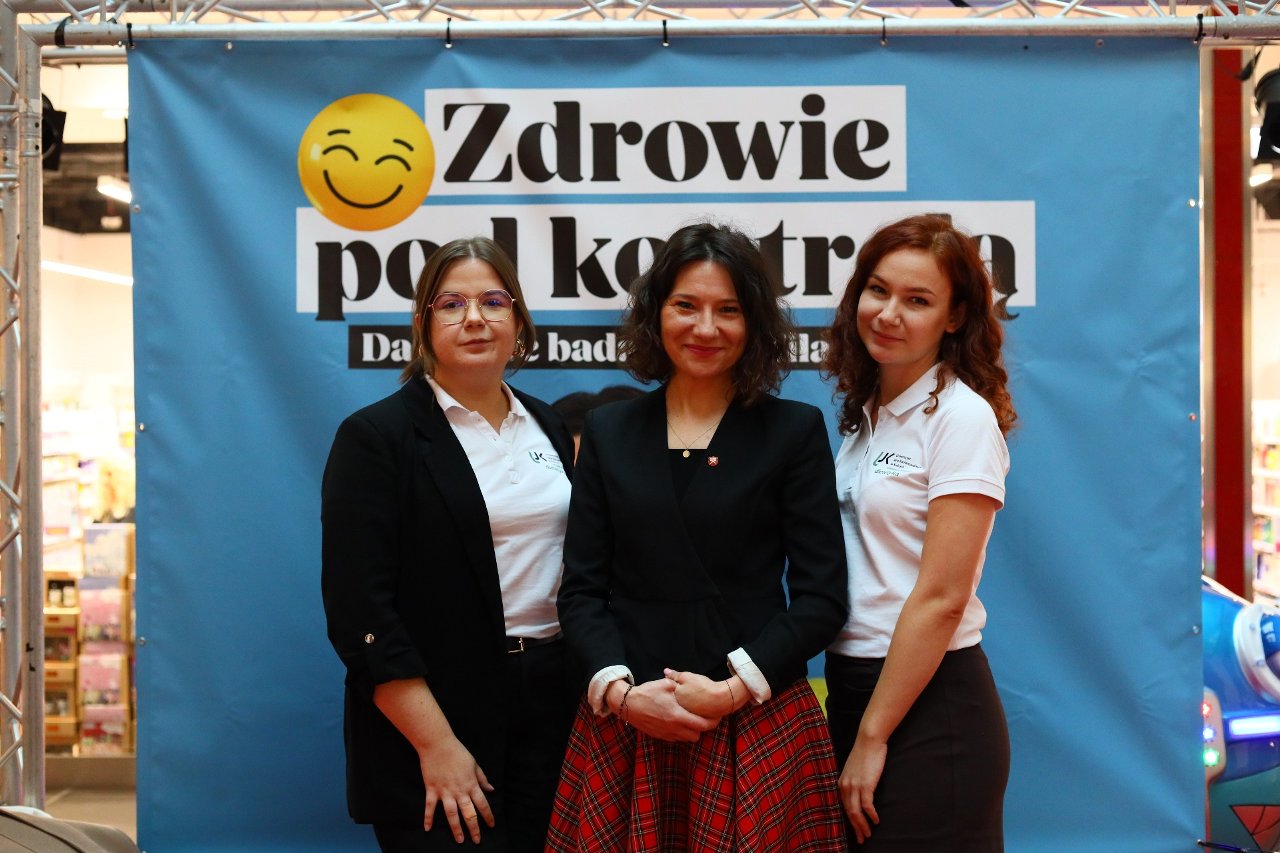 Obraz przedstawia trzy osoby stojące przed niebieskim tłem z banerem zawierającym tekst i duże żółte emoji. Osoby uśmiechają się i stoją blisko siebie pod metalową konstrukcją widoczną u góry kadru. Sceneria wygląda na wnętrze miejsca publicznego lub przestrzeni wydarzeniowej.