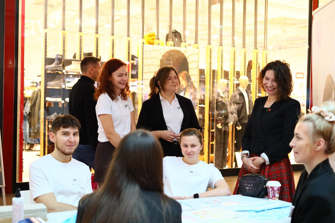 Obraz przedstawia grupę osób w centrum handlowym, skupionych wokół stołu z dokumentami i napojem. Przy stole siedzą dwie osoby, a za nimi stoją kolejne, rozmawiając ze sobą. Obok znajduje się jeszcze jedna osoba obserwująca sytuację. W tle widać przeszkloną ścianę z ekspozycją odzieży.