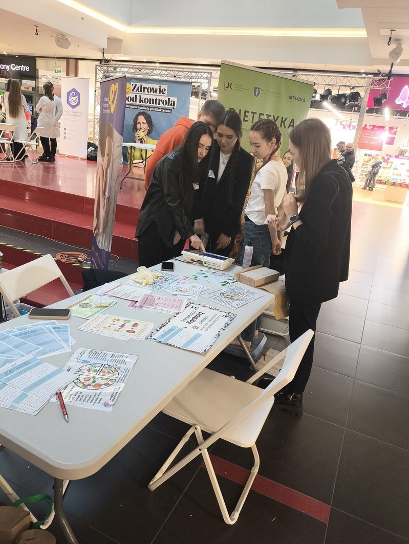 Obraz przedstawia grupę osób w centrum handlowym, zgromadzonych wokół stołu z ulotkami i materiałami informacyjnymi. Po prawej stronie kilka osób przegląda dostępne materiały. W tle widoczne są banery promocyjne oraz sceniczne tło. Za nimi znajdują się sklepy i przechodnie. Podłoga z połyskiem odbija światła z góry.