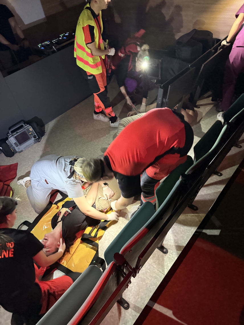 Na obrazie widać wnętrze budynku, prawdopodobnie sali z miejscami do siedzenia. Kilka osób w odzieży służbowej zajmuje się osobą leżącą na noszach. Jedna osoba klęczy obok, inne stoją w pobliżu. Na podłodze znajduje się sprzęt, a scena jest słabo oświetlona, z dodatkowym światłem skierowanym na środek wydarzeń.