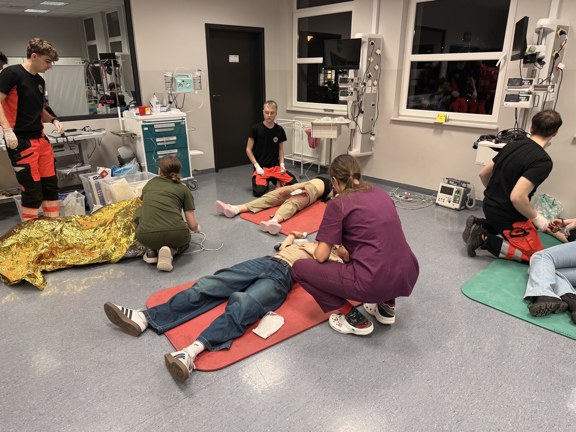 Na obrazie widać salę, w której kilka osób wykonuje czynności przy fantomach leżących na matach. W pomieszczeniu znajdują się elementy wyposażenia oraz wózek ze sprzętem. Część osób pracuje na podłodze, inne stoją obok stołu. Wnętrze jest jasne i uporządkowane, z widocznymi oknami po jednej stronie.