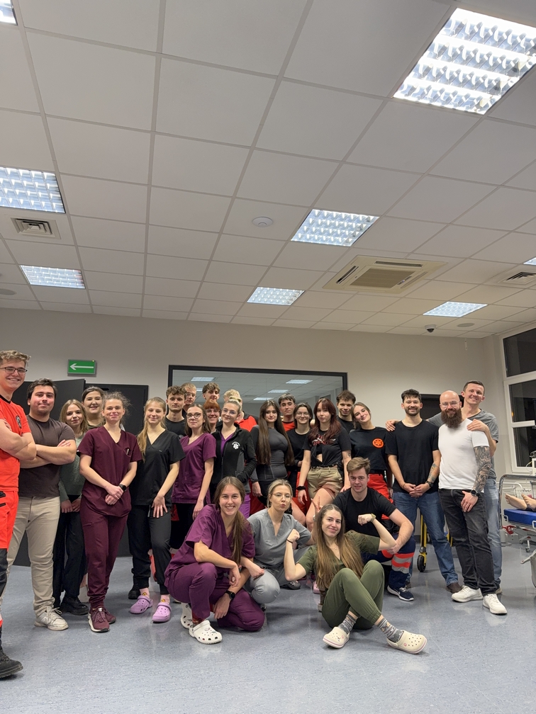 Na obrazie widać liczną grupę osób zgromadzonych w jasno oświetlonym pomieszczeniu. Część osób stoi, inne siedzą lub kucają na podłodze. Uczestnicy mają różnorodny strój, w tym odzież codzienną oraz uniformy. W tle widoczne jest przejście oraz przeszklona powierzchnia odbijająca światło z sufitu.