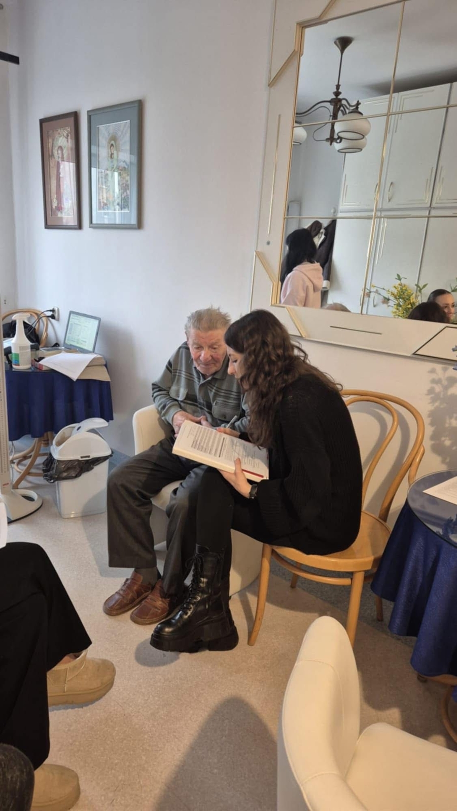 Starszy mężczyzna i młoda kobieta siedzą blisko siebie w jasnym pomieszczeniu i rozmawiają, wspólnie przeglądając książkę lub dokument. Obok znajduje się mały stolik z laptopem i papierami. Wnętrze zawiera lustro odbijające część wyposażenia, oświetlenie oraz obrazy na ścianie.