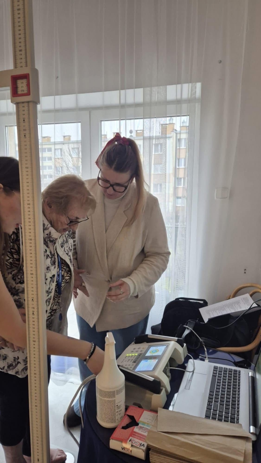 Trzy osoby stoją przy stole z urządzeniami i dokumentami. Dwie z nich wspólnie oglądają kartkę papieru, a trzecia zajmuje się rzeczami leżącymi na stole. Na blacie widać m.in. laptop i butelkę. W tle znajduje się duże okno z widokiem na budynki mieszkalne.