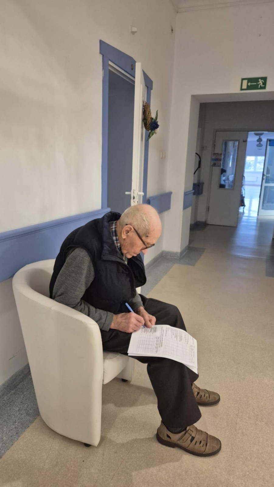 Osoba siedzi na krześle przy ścianie i wypełnia dokument, opierając kartkę na kolanie. W tle widać korytarz z otwartymi drzwiami prowadzącymi do innego pomieszczenia oraz oznaczenie wyjścia ewakuacyjnego.