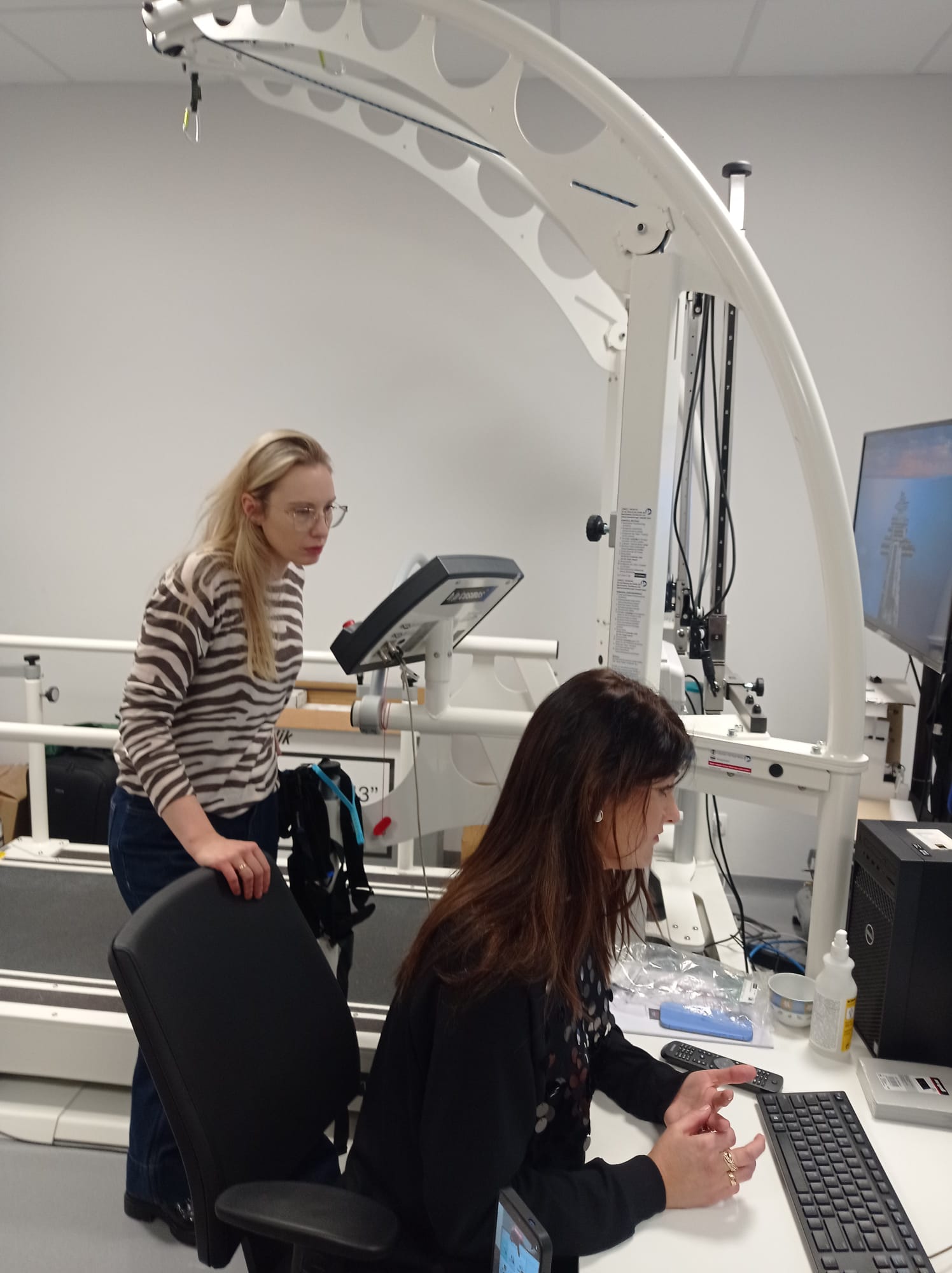 Na obrazie widać dwie osoby w laboratorium lub gabinecie. Skupiają się na specjalistycznym urządzeniu medycznym lub treningowym, podobnym do bieżni. Jedna osoba analizuje panel urządzenia, a druga pracuje przy komputerze.