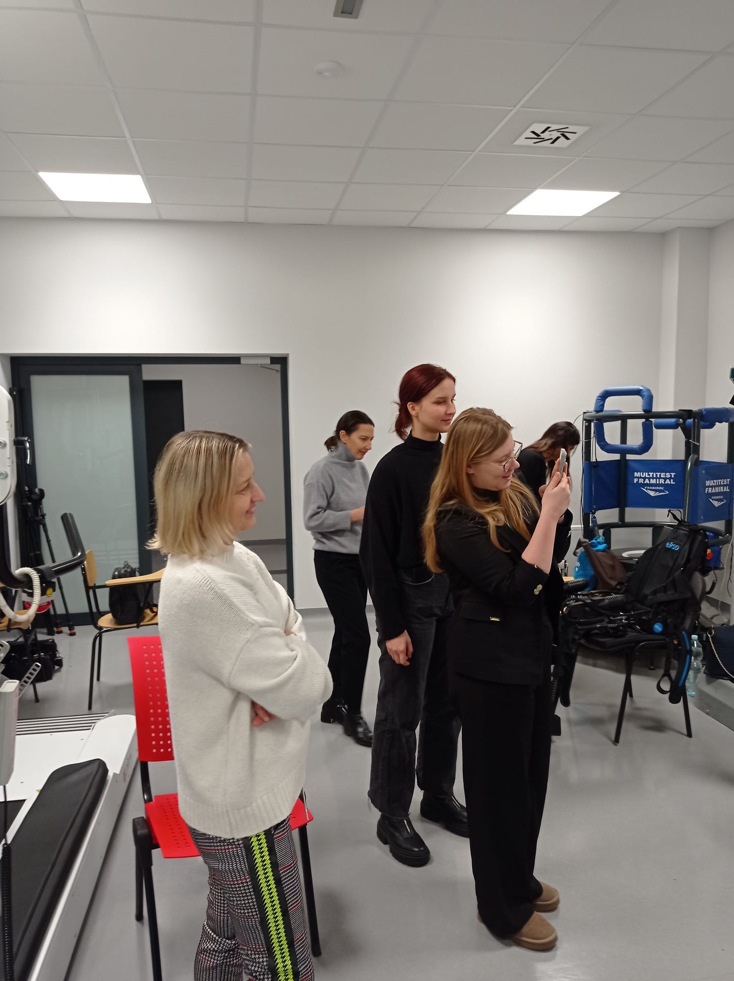 Na obrazie widać cztery osoby stojące w jasnym pomieszczeniu. Część z nich znajduje się na środku sali, jedna osoba korzysta z telefonu, a inna może robić zdjęcie. Po prawej stronie widoczny jest specjalistyczny sprzęt z oznaczeniami „MULTITEST” i „FRAMIRAL”. Pomieszczenie jest przestronne i dobrze oświetlone.