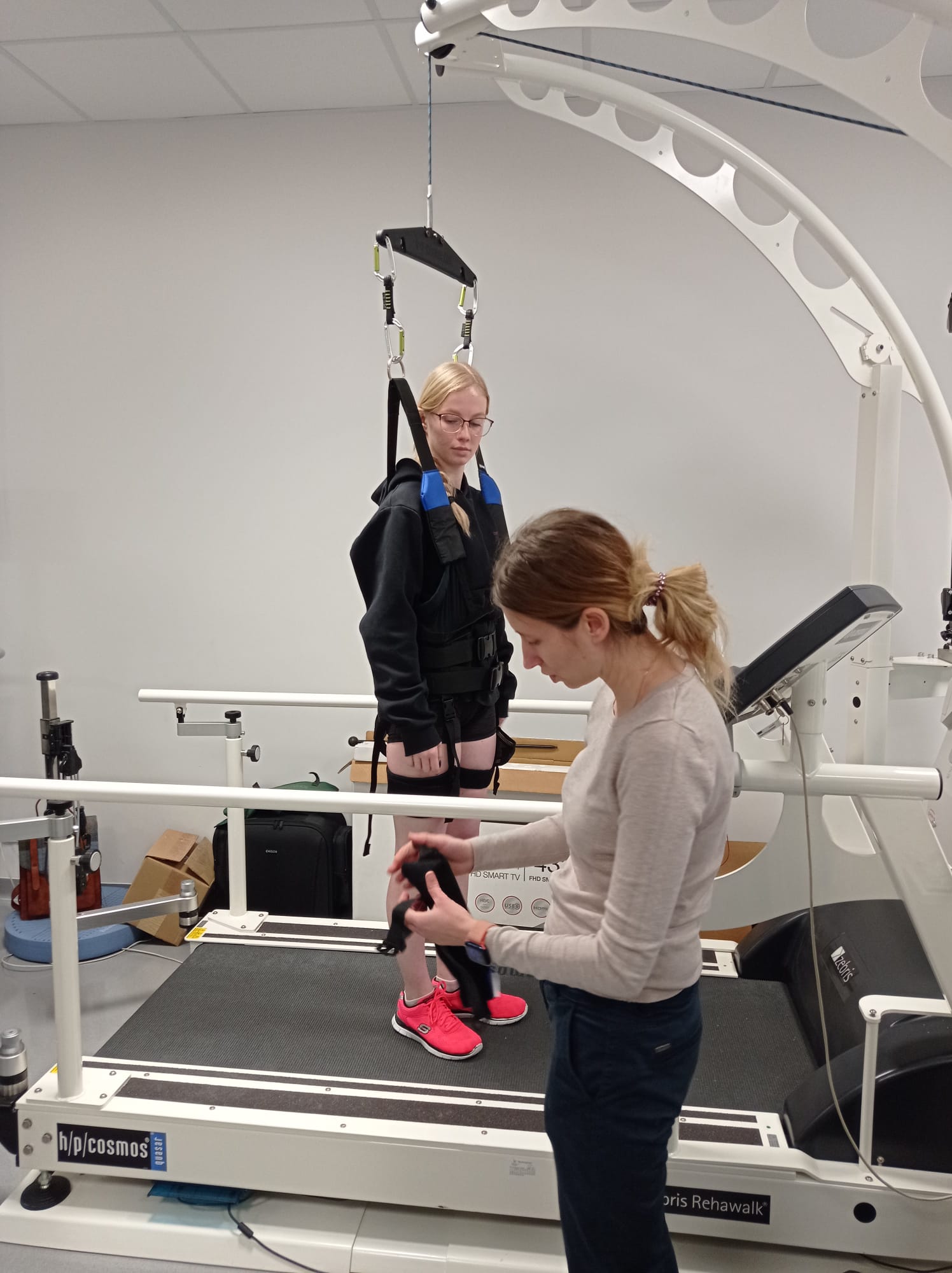 Na obrazie widać laboratorium lub salę rehabilitacyjną. Jedna osoba ćwiczy na bieżni z systemem podwieszenia przymocowanym do konstrukcji sufitowej, a druga obsługuje lub reguluje element urządzenia. W pomieszczeniu znajdują się dodatkowe przedmioty oraz sprzęt, co sugeruje środowisko badawcze lub terapeutyczne.