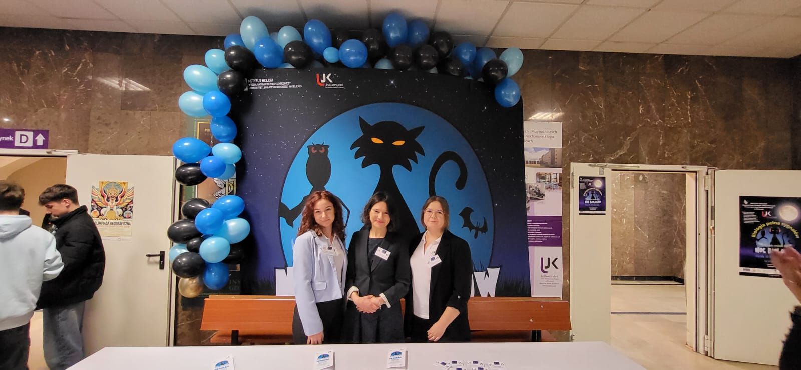 Obraz przedstawia wydarzenie w pomieszczeniu z dużym dekoracyjnym tłem z motywem nocnym, zawierającym ilustracje zwierząt na tle nieba. Nad tłem znajduje się dekoracja z balonów. Na pierwszym planie stoją uczestnicy wydarzenia, pozujący do zdjęcia. W tle widoczne jest wejście do korytarza oraz plakaty umieszczone na ścianach i drzwiach.